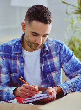 Taşınan bir adam boş bir dairede oturuyor, Kutuların arasında, eşyaların listesini kontrol ediyor.