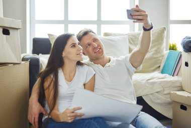 Genç çift yeni evine taşınıyor, yerde oturuyor ve selfie çekiyor.