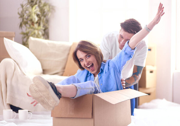 Couple having fun laughing moving into new home, young woman riding sitting in cardboard box while man pushing it