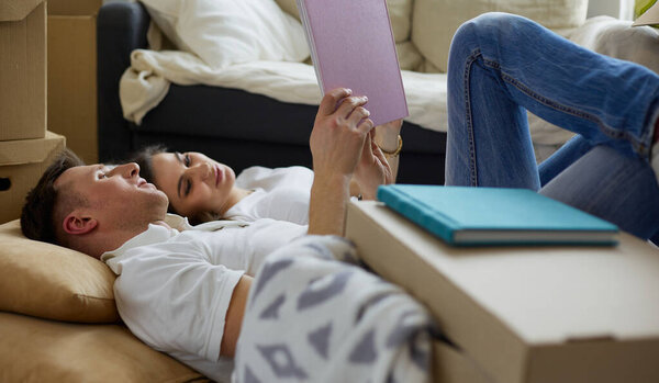 Cute couple unpacking cardboard boxes in their new home, lying on the floor and looking at a family album