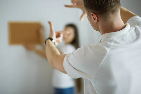 Vrouw helpt haar partner om schilderij te hangen aan de muur van hun nieuwe huis, ze is framing de foto met haar vingers — Stockfoto