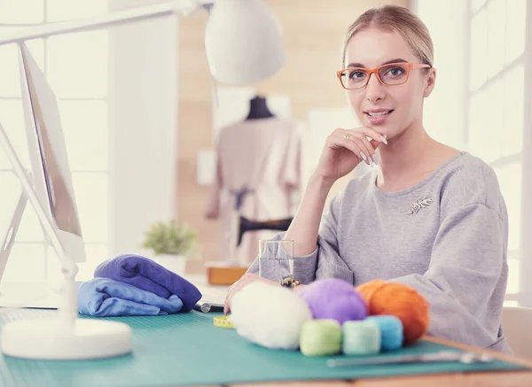 Designer de moda feliz sorrindo, sentado em estúdio de escritório moderno com materiais, tecido, fio, têxtil, laptop na mesa. — Fotografia de Stock