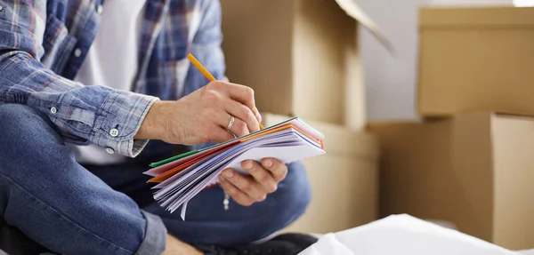 A moving man sitting on the floor in empty apartment, Among the Boxes, Checking the List of Things