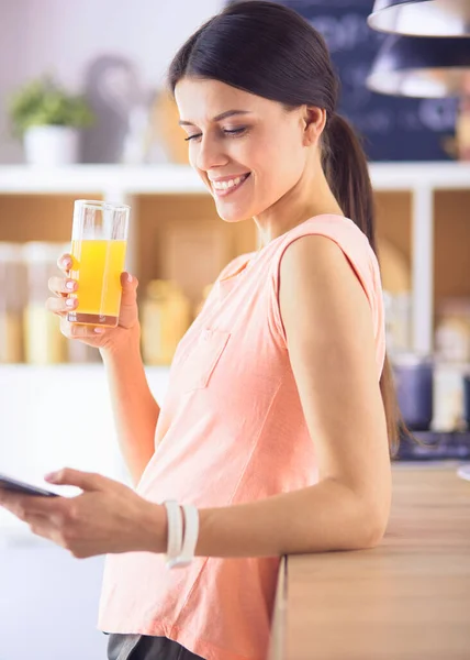 Усміхнена красива жінка дивиться на мобільний телефон і тримає склянку апельсинового соку під час сніданку на кухні . — стокове фото