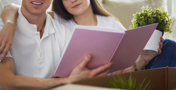 Cute couple unpacking cardboard boxes in their new home, sitting on the floor and looking at a family album — Stock Photo, Image