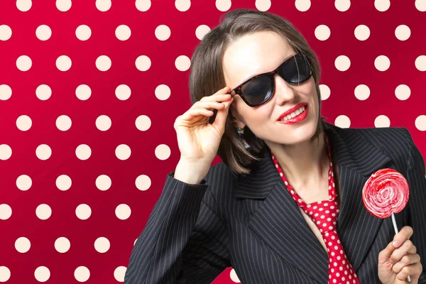 Beautiful woman wearing sunglasses eating candy lolippop over bright red polka dots background — Stock Photo, Image