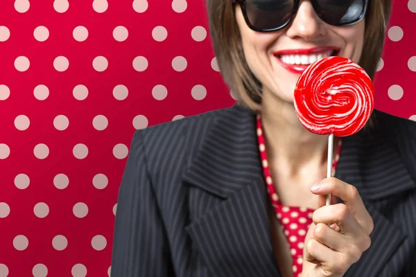 Beautiful woman wearing sunglasses eating candy lolippop over bright red polka dots background — Stock Photo, Image