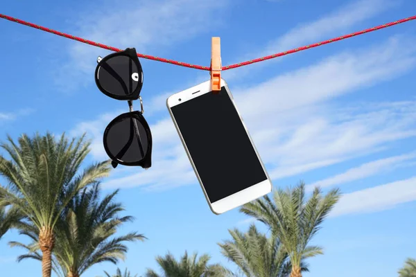 Téléphone et lunettes de soleil suspendues à une corde avec la paume et le ciel sur le fond — Photo