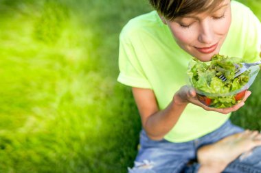 Mutlu gülümseyen kız açık havada yeşil çimenlerin üzerinde oturan sebze salata yemek. Sağlıklı yaşam, kopya alanı