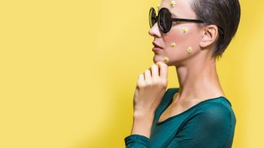 Güzel bir kadın sarıya yakın stüdyoda güneş gözlüğü takıyor ve yüzünde çiçekler var.