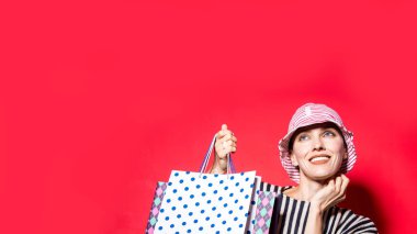 Parlak kırmızı arka planda alışveriş torbaları olan, çizgili şapka ve gömlek giyen mutlu bir alışveriş kadını.