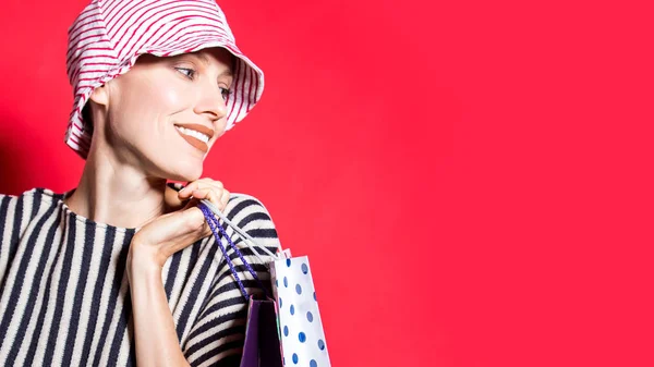 幸せなショッピング女性でショッピングバッグ以上明るい赤の背景にストライプの服の帽子とシャツを身に着けて — ストック写真
