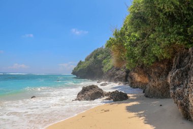Pantai Gunung Payung plajı, güzel altın kumsallı, Bali. Dalgalar kıyıdaki kayalık kayalıklara çarptıkça turkuaz mavi su deniz köpüğüne dönüşür. Mavi gökyüzü güneşli. Turist doğası.