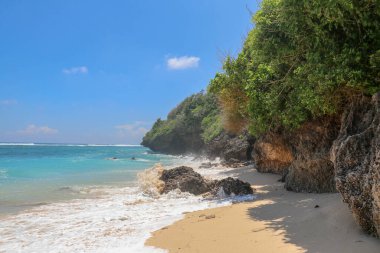 Pantai Gunung Payung plajı, güzel altın kumsallı, Bali. Dalgalar kıyıdaki kayalık kayalıklara çarptıkça turkuaz mavi su deniz köpüğüne dönüşür. Mavi gökyüzü güneşli. Turist doğası.