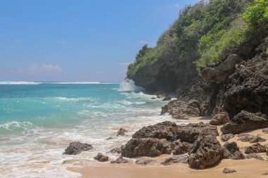 Pantai Gunung Payung plajı, güzel altın kumsallı, Bali. Dalgalar kıyıdaki kayalık kayalıklara çarptıkça turkuaz mavi su deniz köpüğüne dönüşür. Mavi gökyüzü güneşli. Turist doğası.