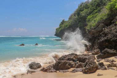 Pantai Gunung Payung plajı, güzel altın kumsallı, Bali. Dalgalar kıyıdaki kayalık kayalıklara çarptıkça turkuaz mavi su deniz köpüğüne dönüşür. Mavi gökyüzü güneşli. Turist doğası.