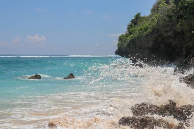 Pantai Gunung Payung plajı, güzel altın kumsallı, Bali. Dalgalar kıyıdaki kayalık kayalıklara çarptıkça turkuaz mavi su deniz köpüğüne dönüşür. Mavi gökyüzü güneşli. Turist doğası.