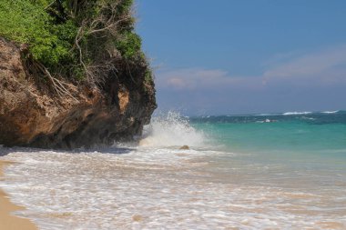 Pantai Gunung Payung plajı, güzel altın kumsallı, Bali. Dalgalar kıyıdaki kayalık kayalıklara çarptıkça turkuaz mavi su deniz köpüğüne dönüşür. Mavi gökyüzü güneşli. Turist doğası.
