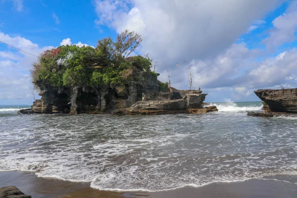 Waves shatter on a cliff at the top of which is the Hindu temple of Tanah Lot. Temple built on a rock in the sea off the coast of Bali island, Indonesia. Lush tropical vegetation on top of a cliff.