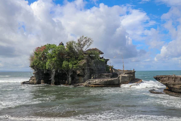 Waves shatter on a cliff at the top of which is the Hindu temple of Tanah Lot. Temple built on a rock in the sea off the coast of Bali island, Indonesia. Lush tropical vegetation on top of a cliff.