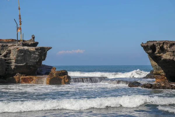 Wave crashing onshore at low tide in Bali, Indonesia. Water cascading over stone, feeding tidal pool. Wave breaking on rock during low tide near Tanah Lot. Deep blue ocean and sky in the background.