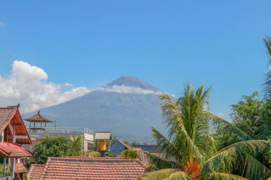 Arka planda palmiye ağaçları ve görkemli yanardağı olan tropik bir cennet. Volkan kraterinin etrafında beyaz bulutlar. Evin çatısından manzaraya bak..