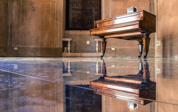 Old rustic piano on stands with wheels in empty room with polished glass look floor making reflection of piano and whole room symmetrical