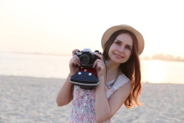 Genç, güzel esmer, gülümseyen bir kız elinde kamerayla kıyıda duruyor.