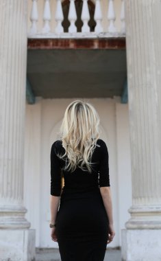 Güzel bir vücudu ve uzun sarı saçları olan genç ve güzel bir kadın beyaz bir binanın arka planında duruyor.