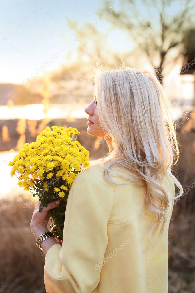 beautiful blonde in a yellow coat and with a bouquet of yellow chrysanthemums stands on the river
