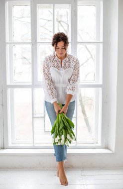 Genç Avrupalı siyah saçlı kız beyaz bir stüdyoda pencerenin yanında duruyor ve elinde bir buket beyaz lale tutuyor.