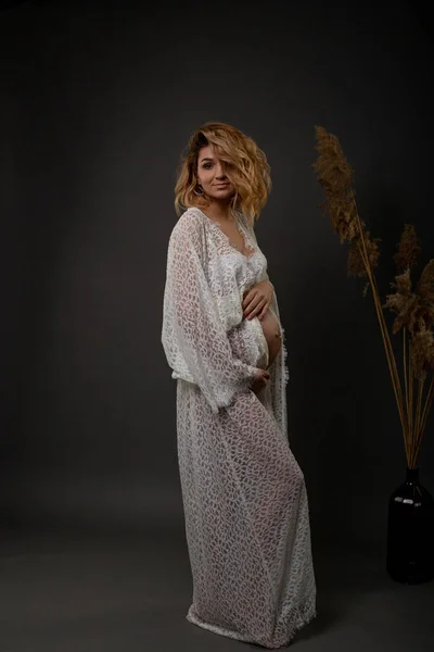 年轻的怀孕的金发女郎站在黑暗的背景下 穿着长长的白色长袍 — 图库照片