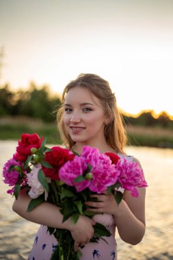 Elinde bir buket kırmızı ve pembe şakayıkla gülümseyen kız güzel bir arka planda duruyor.