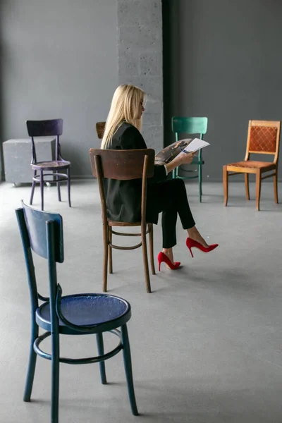 young blonde sits on a chair in a half-empty room and leafs through a magazine