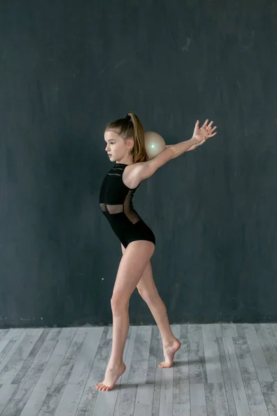 charming little gymnast standing exercises with the ball against the background of the dark gray wall