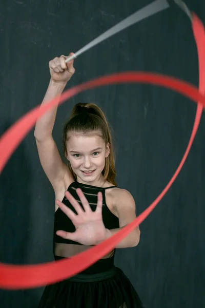smiling little gymnast standing facing the camera performs an exercise with a bright red ribbon on a dark gray background