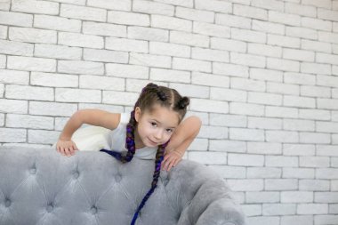 Uzun ince örgülü güzel bir kız, gri bir kanepenin yanında, tuğla bir duvara karşı oynuyor.