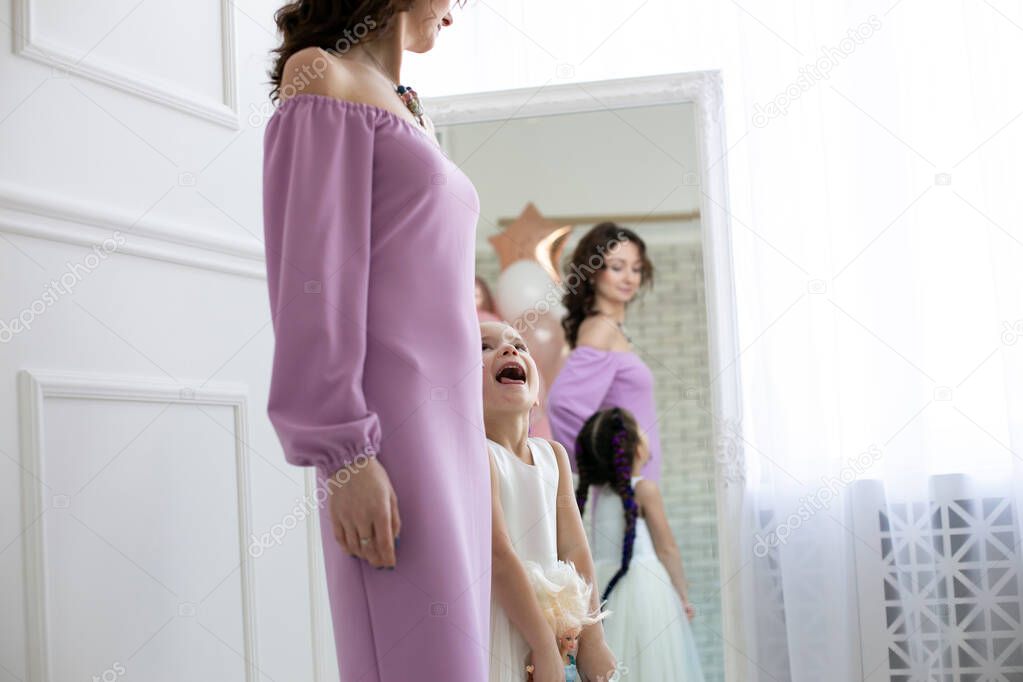 young slim woman in a purple dress and her five year old daughter in a white dress are standing in a white studio near the mirror