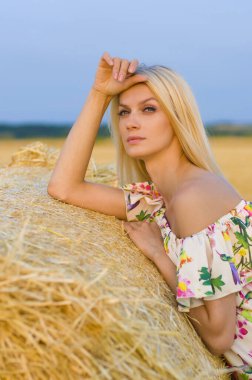 Çıplak omuzlu, çok renkli bir elbise giymiş üzgün bir sarışın samanlığın yanında duruyor tarlanın ve gökyüzünün arka planında.