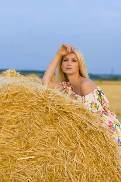 美丽的金发姑娘光着肩膀 穿着五颜六色的衣服 站在田野和天空的衬托下的草堆旁 — 图库照片