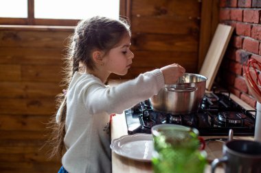 Kız, pencere kenarındaki bir kır evinin mutfağındaki ocakta yemek yapmayı öğreniyor.