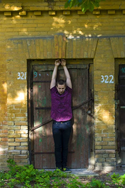 年轻伤心的小伙子站在一座靠近木门的旧砖楼的后面 双手高举 — 图库照片