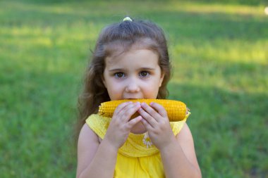 Uzun kıvırcık saçlı komik küçük kız yeşil çimlerin üzerinde duruyor ve mısır yemekten hoşlanıyor.