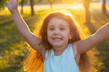 Uzun kıvırcık saçlı neşeli küçük kız kollarını açmış, yaz parkının yeşil çimlerinde koşuyor.