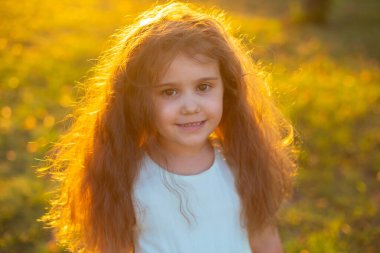 Gün batımında yeşil çimlerin üzerinde duran uzun kıvırcık saçlı tatlı esmer kız kameraya gülümsüyor.