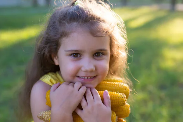 長い巻き毛の少女は手にトウモロコシの腕を持つ緑の芝生の夏の公園に立っています — ストック写真