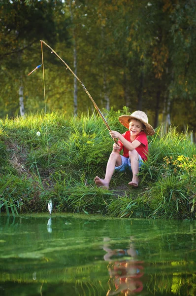 一个光着脚的快乐男孩坐在河岸上 手里拿着钓竿 钓着鱼 — 图库照片