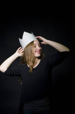 Uzun saçlı güzel sarışın ayakta duruyor ve gülümsüyor. Başındaki karton tacı düzeltiyor.