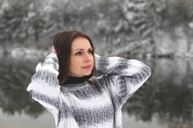 Gri kazaklı güzel, uzun saçlı esmer. Kış ormanı nehrinin karlı kıyısında elleri başının arkasında duruyor.