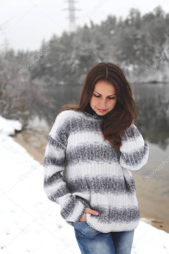 pretty long-haired brunette in a gray sweater and blue jeans stands on the snowy shore of a winter forest river with her head down